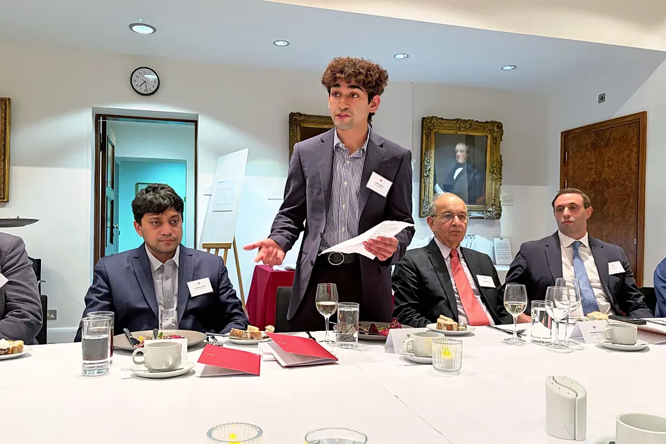 Ashwin Telang stands at the dinner table holding notes and gestures as he presents, with seated guests on either side.