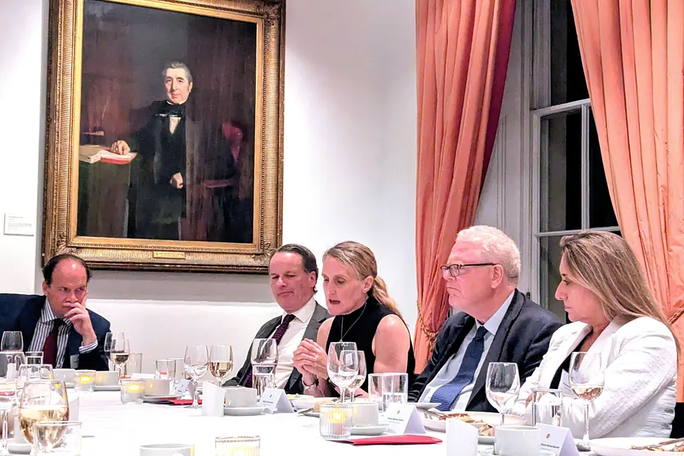 A guest seated at the table speaks while others listen, beneath a large gilt-framed portrait.